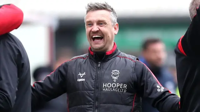 Michael Skubala, wearing a black Lincoln with red trim, has a beaming smile on his face after the Imps beat AFC Wimbledon 1-0