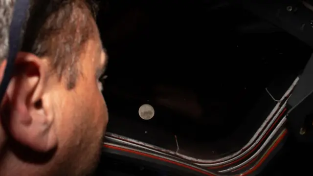 Artemis II commander and Nasa astronaut Reid Wiseman looking out the window of the spacecraft to look at the Moon which is light and dark grey surrounded by pitch-black sky