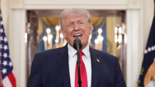Donald Trump in a dark suit and red tie speaking into a microphone.