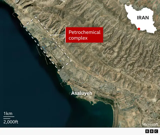 A satellite map of the coastal city of Asaluyeh in southern Iran, showing a large petrochemical complex along the shoreline. The complex is highlighted with a red label and outlined by dashed white lines. Mountains lie to the northeast, and the coastline runs north–south along the left side of the image. A small inset map of Iran in the top-right corner marks the location of Asaluyeh with a red dot. A scale bar indicates 1 km (2,000 ft).