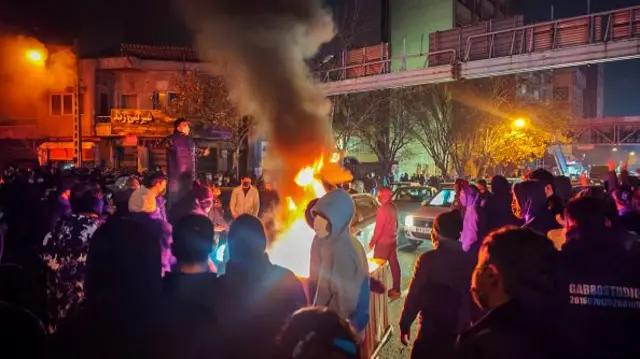 A fire is lit and people gather round at night time in a Tehran street