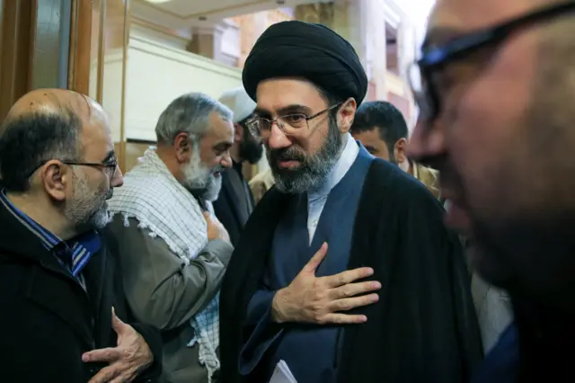 Mojtaba Khamenei wearing a black turban, black cloak and grey suit as he walks among other men with his right hand over his heart