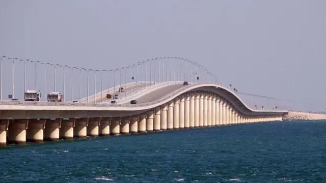 A large curved concrete bridge is shown crossing a stretch of water, with cars and trucks driving over it.
