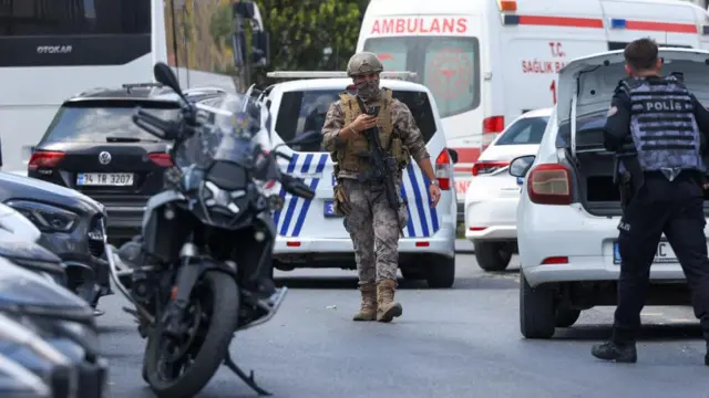 An officer wearing camouflage holds up a phone as he walks through a car park. A police officer can be seen standing next to the open boot of a car