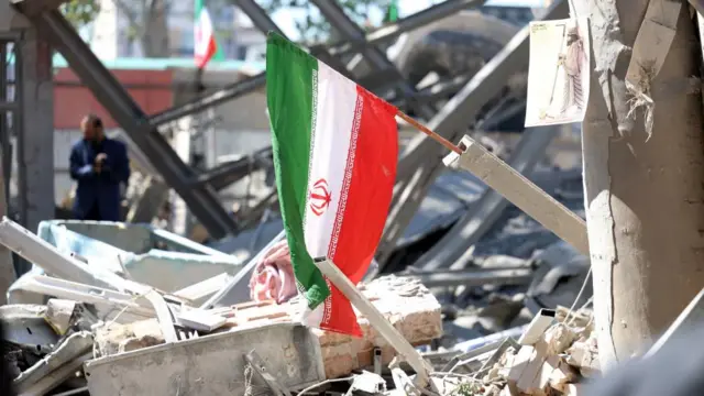 हमले की चपेट में आई एक इमारत के मलबे और मलबे में एक ईरानी राष्ट्रीय ध्वज फंस गया। पृष्ठभूमि में लोगों को मलबे को देखते हुए देखा जा सकता है