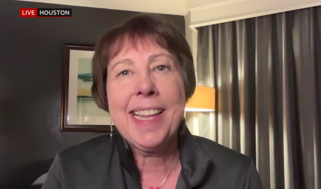 Dr Nicola Fox, a white lady with brown hair, sat in a room with curtains drawn and a lamp on, smiling at the camera.