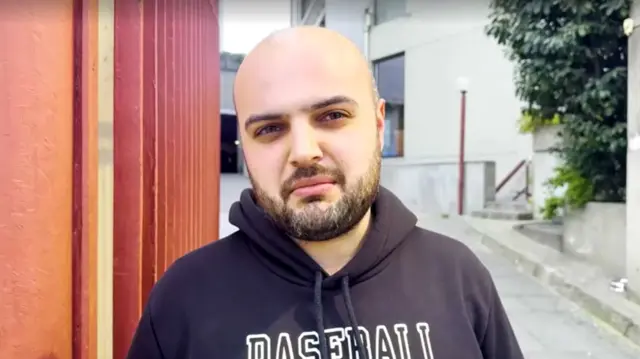 Man in hooded jumper speaks to camera in middle of a quiet street