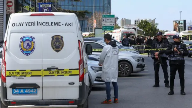A man in a white jacket stands close to a white van. Several officers stand holding guns next to some police tape