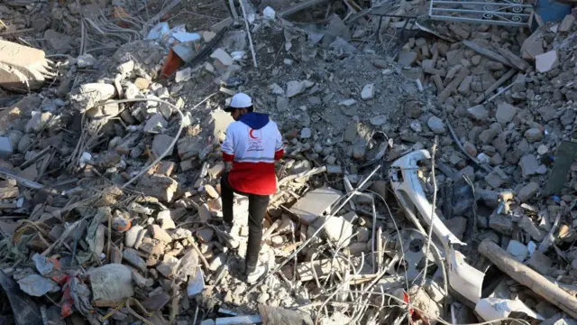 'ईरानी रेड क्रिसेंट' लोगो, अर्धचंद्राकार हाई विज़ जैकेट पहने एक व्यक्ति मलबे के विशाल ढेर के बीच से चलता हुआ