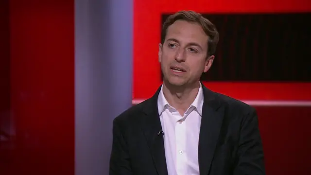 Gideon Falter is shown in a white shirt and black blazer in the BBC News studio against a red background.