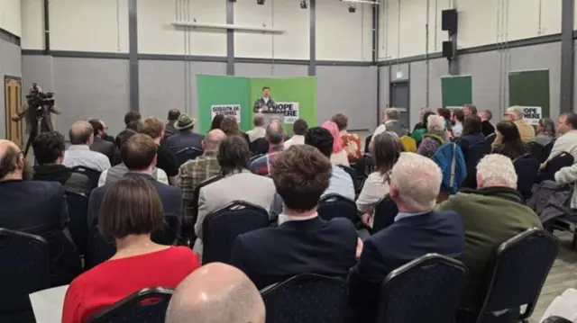 Behind view of the audience sat looking at a young man on stage.