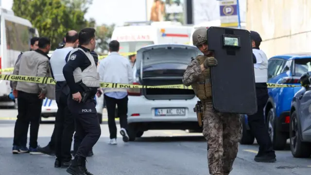 A soldier carries a riot shield away from police tape as several officers wearing vests over their uniforms stand nearby. A car with its boot open can be seen in front of the group