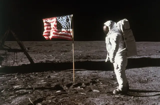 Astronaut Buzz Aldrin in a white outfit and large helmet stands beside an American flag placed on the moon during Apollo 11