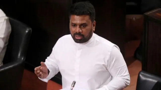 President Anura Kumara Dissanayake speaks behind a microphone in parliament. He is standing, and wearing a long-sleeved white shirt.