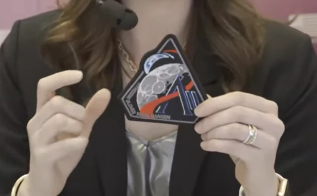 A triangular patch showing stylised representations of the Moon in front of the Earth, with the AII logo of Nasa's Artemis II mission is held by a person.