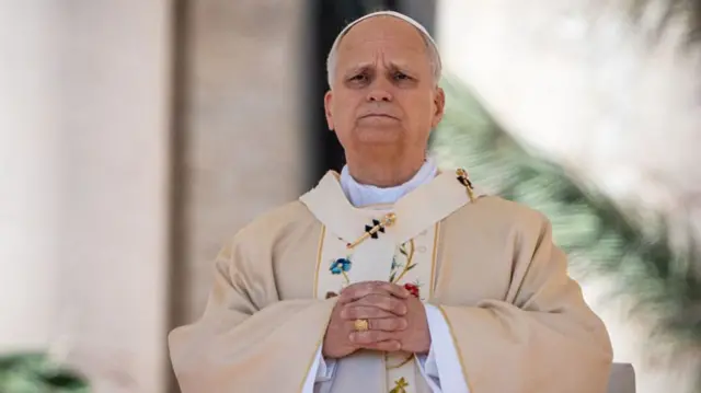 Pope Leo stands in a cream-colored robe with his hands folded over his chest