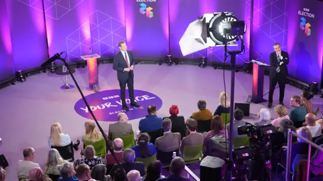 Audience members are sat in chairs on rows looking at a man in a suit standing up/