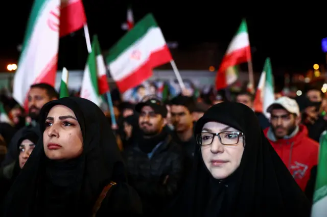 Two women stand in a crowd waving Iranian flags. They both wear black hijabs and one has rectangular glasses on.