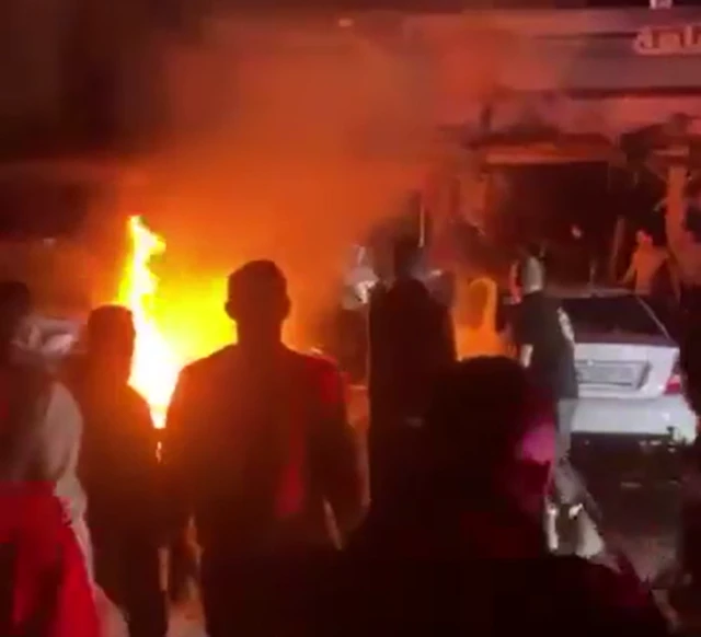 A screenshot from a video of around 5 people looking at a large fire and smoke outside a damaged building at night