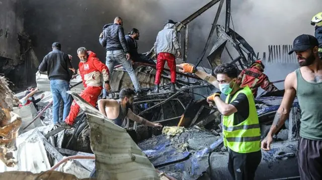 A group of men climb over the smouldering wreckage of a building. A man in a safety vest and mask appears to be directing their movements.