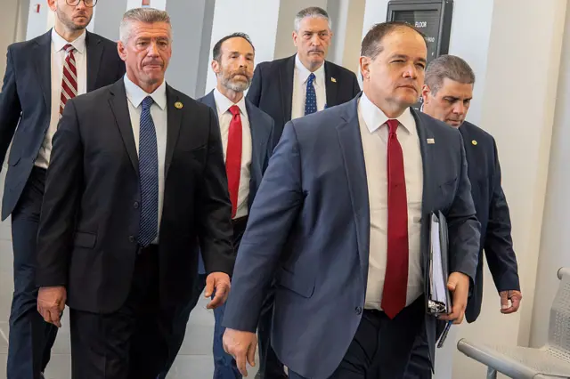 Six men in grey suits and red or blue ties walk down a hallway
