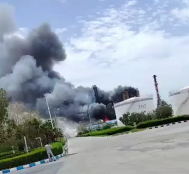 Smoke billows from an oil refinery building which is on fire, two storage tanks are visible nearby