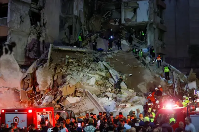 Rescue crews search rubble. A large crowd is gathered