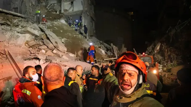 Emergency workers wearing full body equipment and some with torches on their head are climbing through a huge pound of rubble in darkness. One is turning towards the camera, frowning and shouting