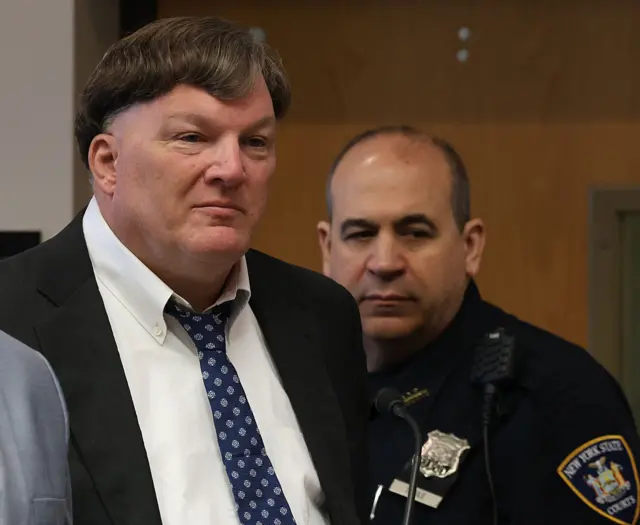 A man in a suit and blue patterned tie looks off to the right as a security guard stand behind him