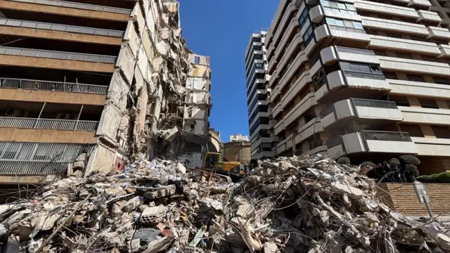 A picture of rubble piled up in between two large residential looking buildings in Beirut. There's a digger on top of the rubble in the background.
