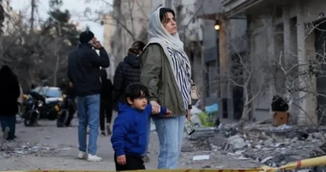 A woman and her child look at the aftermath of an air strike in Tehran. Debris surrounds them while the look on next to a cordon