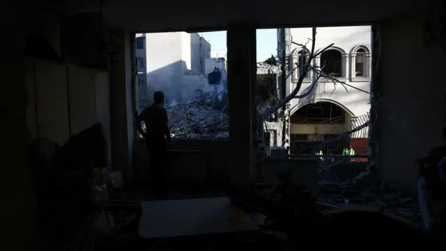 A man stands in a dark room and stares out at a pile of rubble