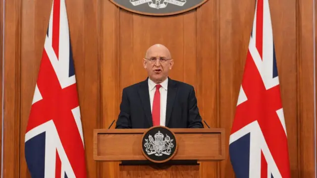 John Healey delivering his statement in front of two British flags