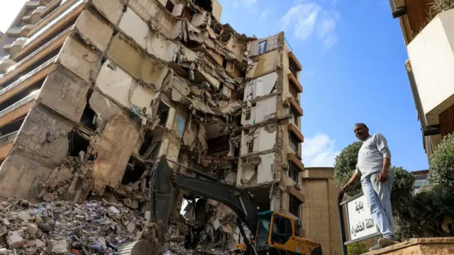 A man stands outside a damaged building in Lebanon. There are large piles of rubble and debris and the building is clearly exposed