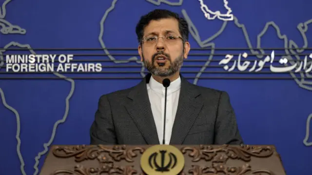 Saeed Khatibzadeh speaks behind a wooden lectern in front of a large map of the world.