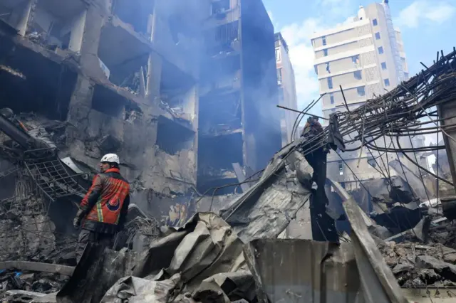 A man in an orange safety jacket and helmet stands on the smoking ruins of a building hit by a strike.