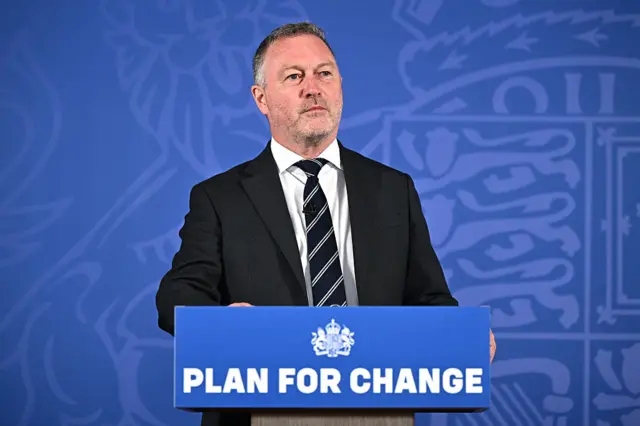 Steve Reed, Secretary of State for Housing, Communities and Local Government of the United Kingdom, delivers a speech at a blue podium with the words 'Plan For Change' on it. He is wearing a suit and tie.
