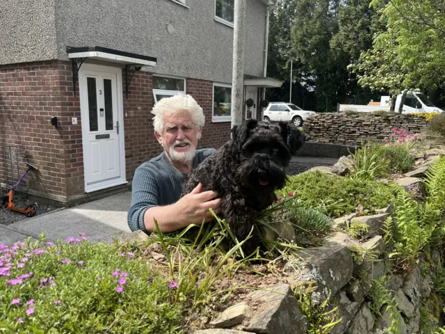 A man with a beard and a dog sitting on a hedge in front of a house