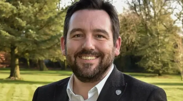 David Baines, a man in a black blazer and white shirt, smiles into the camera, standing in a field with grass and trees.
