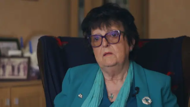 Teresa Russell sitting on an armchair wearing glasses, a blue blazer and scarf.