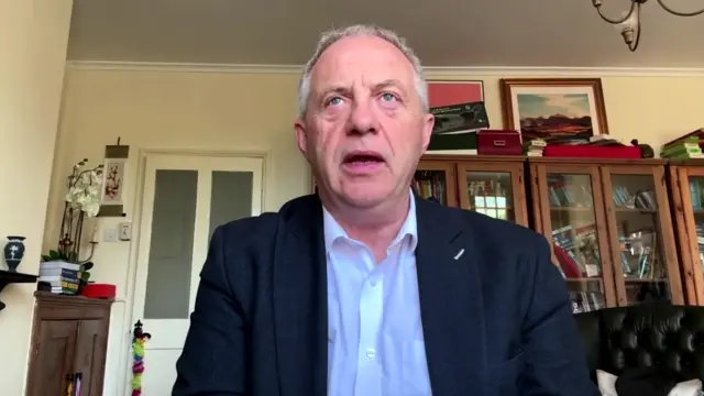 Lord Mann speaks from what appears to be a room in his house while wearing a white shirt and dark blazer