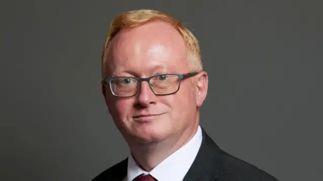 A picture of MP Luke Akehurst. He was giner hair and glasses, wearing a suit with a black blazer and red tie.