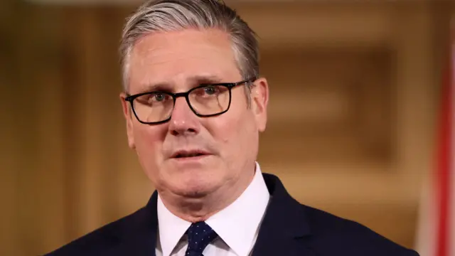 Sir Keir Stamer looking to the right of the camera with a serious expression. He is wearing black framed glasses, a black suit jacket and tie, and a white shirt