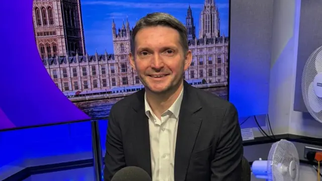 Chris Ward, in a blazer and white shirt, smiles towards the camera, sitting in a radio studio.