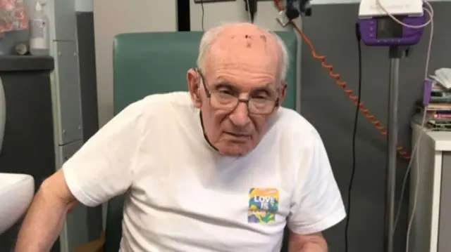 John Russell in the hospital. He is wearing glasses and a white t-shirt.