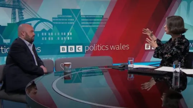 Llyr Powell and Teleri Glyn Jones are sitting across from each other on a table in a TV studio.