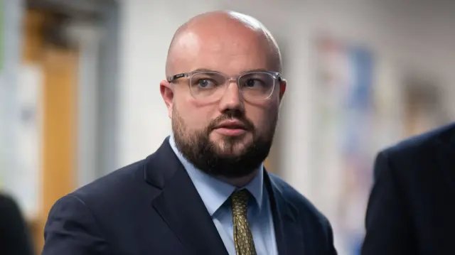 Llyr Powell is looking away from the camera wearing a suit and glasses.