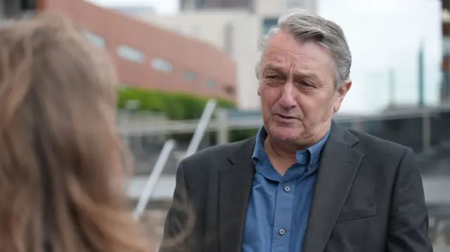 Anthony Slaughter is wearing a suit and is looking at a journalist while speaking to her.