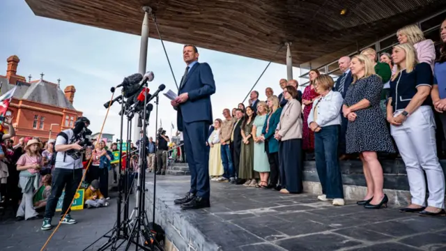 Rhun ap Iorwerth is standing infront of microphones with rows of people standing behind him.