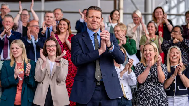 Rhun ap Iorwerth is clapping , with rows of people stood behind him also clapping.